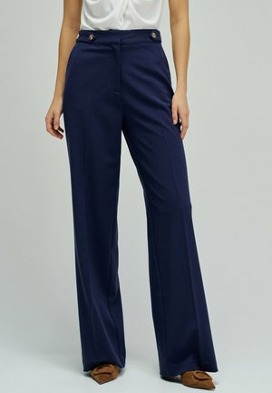 Mujer vistiendo pantalones anchos de talle alto en azul marino con detalles de botones dorados, combinados con zapatos planos marrones de punta puntiaguda.