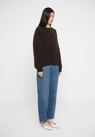 Brun strikket sweater med rund hals, parret med løse blå jeans og spidse hvide hæle, præsenteret mod en simpel baggrund.