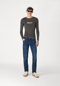 Camisa gris de manga larga con ribete crema y logo, combinada con jeans azules y zapatillas crema con suelas marrones, de pie sobre un fondo liso.