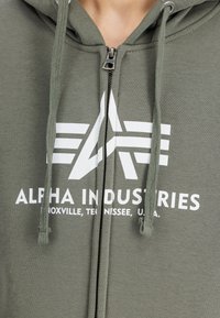 Felpa con zip verde oliva realizzata in misto cotone. Presenta il logo bianco "Alpha Industries" e un cappuccio con cordini in texture a maglia.