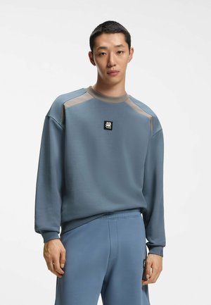 Mężczyzna ubrany w niebiesko-szarą bluzę i dopasowane spodnie, stoi na jednolitym białym tle, zwrócony twarzą do przodu z neutralnym wyrazem.