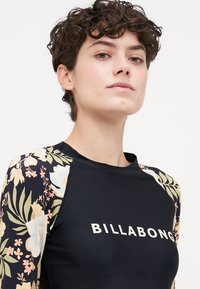 Črna majica z dolgimi rokavi z floralnim vzorcem, s belim napisom "BILLABONG". Narejena iz gladkega, raztegljivega materiala.