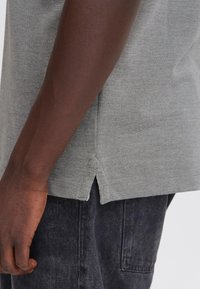 Person mit einem hellgrauen, strukturierten Shirt mit kleinem seitlichen Schlitz und dunkler Hose, linker Arm entspannt an der Seite, vom mittleren Oberkörper abwärts gezeigt.