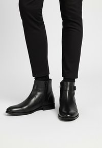 Bottes à cheville en cuir noir avec une finition lisse, un panneau latéral texturé et une fermeture éclair latérale. La semelle est plate et le design est élégant.