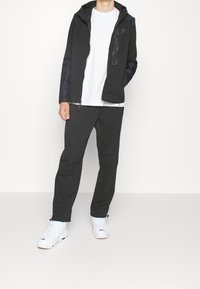 Svart jacka med sidopaneler, huva och dragkedja, i kombination med svarta byxor och vita sneakers. Outfitten har en avslappnad design.