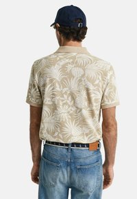 Beige polo shirt med korte ærmer og hvidt tropisk blomstermønster, parret med blå denim jeans og en marineblå kasket. Synlig mærketag på waistband.