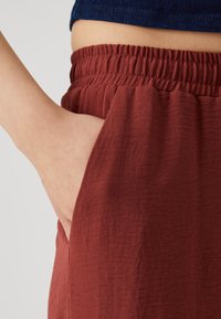 Rust-colored fabric with a crinkled texture; elastic waistband; hand pocket visible on the side; bordering denim top partially seen.