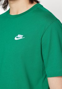 Osoba v zeleném tričku Nike s bílou vyšívanou nášivkou Nike na hrudi, zaměřeno na detaily ramen a krku.