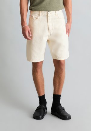 Mężczyzna ubrany w kremowe krótkie spodenki jeansowe, czarne skarpetki, czarne buty wsuwane oraz oliwkowo zieloną koszulę, stojący na tle szarego tła.