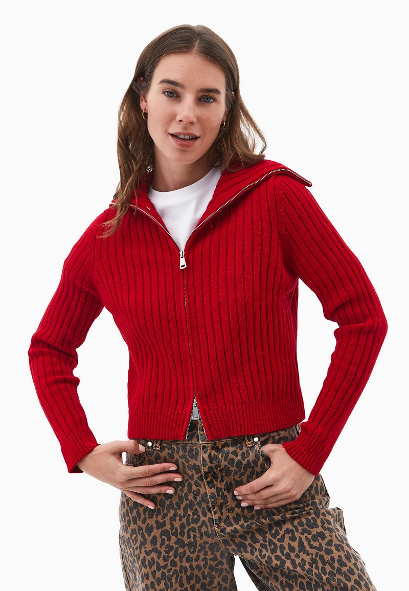 Czerwony sweter z zamkiem błyskawicznym, z wysokim kołnierzem i długimi rękawami, o teksturowym wykończeniu. Noszony na spodniach w panterkę.