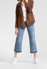 Chaqueta de ante marrón con cremallera frontal, blusa blanca con flores y jeans cortos de tiro bajo en azul claro con dobladillo deshilachado y zapatillas blancas.