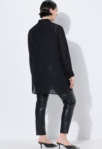 Zwarte chiffon blouse met verticale plooien, lange mouwen en een kraag, gecombineerd met strakke zwarte leren broeken en puntige zwarte hakken.