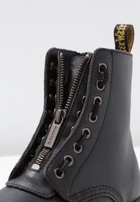 Bota de cuero negra con cremallera parcialmente abierta, centrada entre ojales metálicos y cordones negros, que cuenta con una lengüeta amarilla con la marca.