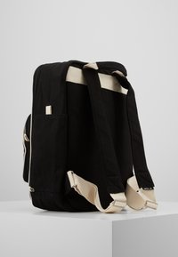 Mochila preta de lona com alças creme. Apresenta dois bolsos laterais e fecho de zíper. Inclui um pequeno rótulo com "MELA" numa das alças.