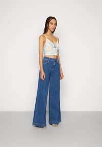 Top floral azul cortado com um nó na frente, combinado com jeans azuis de perna larga. Sandálias de salto transparentes completam o visual.