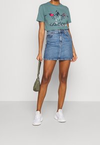 Blå denimminikjol ihop med en teal t-shirt med en blommig Adidas-logotyp. Vita sneakers och en olivgrön handväska fulländar looken.