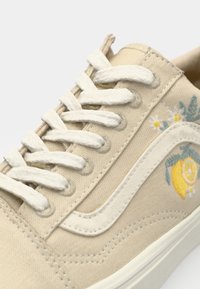 Beige canvas sneaker with white laces, featuring yellow lemon and floral embroidery on side, textured stitching, and rubber sole.