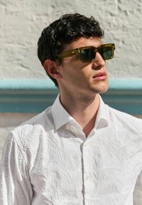 Groene vierkante zonnebril met een doorschijnend olijfgroen montuur; gedragen met een witte geborduurde blouse met bloemmotieven.