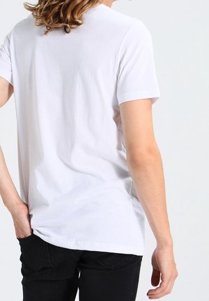 Osoba s vlnitými hnědými vlasy, oblečená v jednoduchém bílém tričku s krátkým rukávem a černých kalhotách, zobrazená zezadu s jednou rukou v kapse.