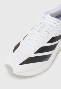 Chaussure de sport blanche avec des rayures diagonales noires, dotée d'une tige texturée, d'un design léger et de lacets blancs. Semelle en caoutchouc lisse.