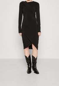 Černé šaty s dlouhým rukávem a diagonálním řasením s asymetrickým spodním lemem. Doplněné špičatými černými koženými botami na silném podpatku.