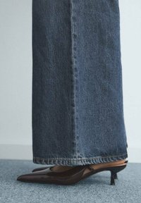 Piernas inferiores en jeans de mezclilla azul largo combinados con zapatos de tacón bajo de punta marrón sobre una alfombra gris.