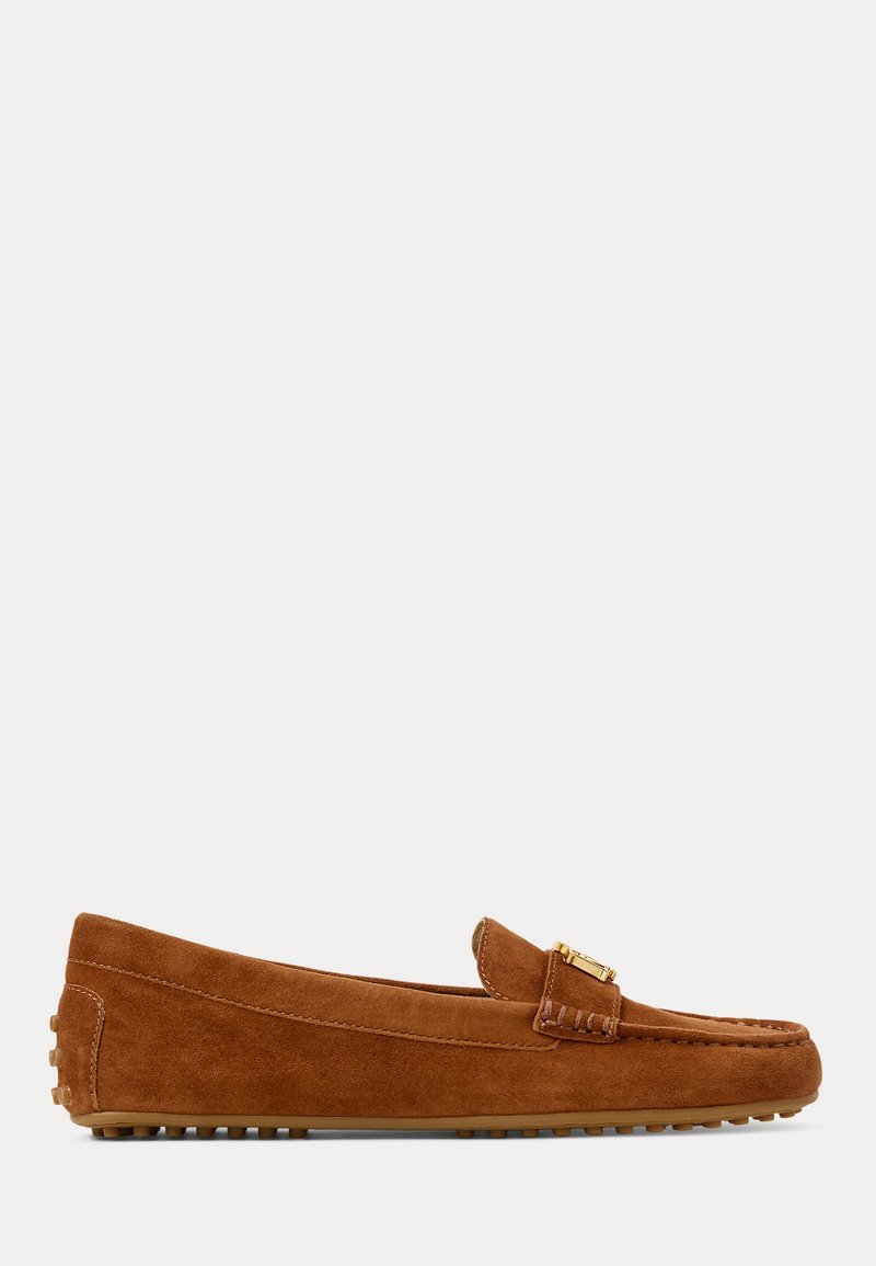 Zapato mocasín marrón de ante con detalle de hebilla dorada y suela de goma con textura de piedrecillas, visto de lado sobre un fondo liso.