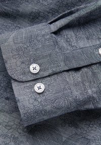 Gros plan sur un poignet de chemise bleue à motifs avec deux boutons blancs et des motifs floraux et feuillus détaillés sur le tissu.