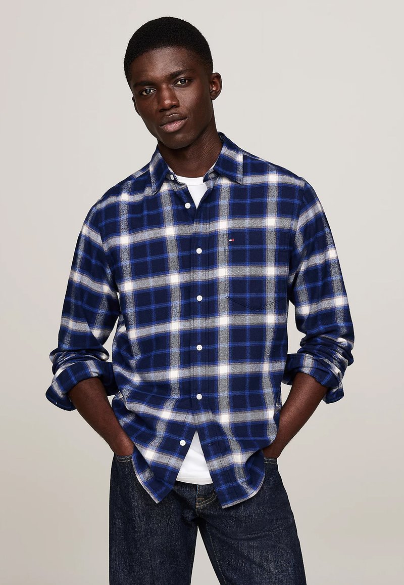 Camicia in flanella a quadri blu navy e bianca con colletto ribattuto, taschino singolo sul petto e maniche arrotolate, indossata sopra una maglietta bianca.