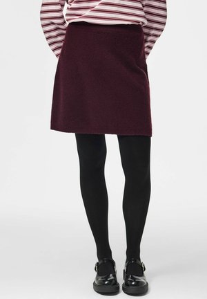Femme portant une jupe bordeaux, des collants noirs, des chaussures Mary Jane noires et un haut à manches longues rayé blanc et bordeaux.