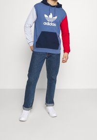 Color-blocked hoodie with navy, light blue, red, and grey sections; features a kangaroo pocket and white adidas logo. Paired with denim jeans.