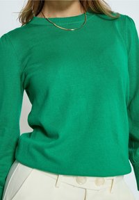 Grüner Strickpullover mit rundem Halsausschnitt, gerippten Bündchen und glatter Textur, kombiniert mit maßgeschneiderten off-weißen Hosen mit großen Knöpfen.