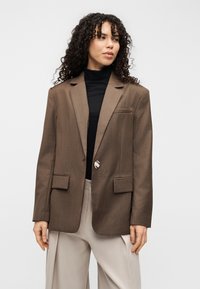 Καφέ κομψό σακάκι με ανασηκωμένες πέτρες, μία κουμπιά κλεισίματος και δύο μπροστινές τσέπες. Φορεμένο πάνω από μαύρο ζιβάγκο, με ανοιχτόχρωμο παντελόνι.