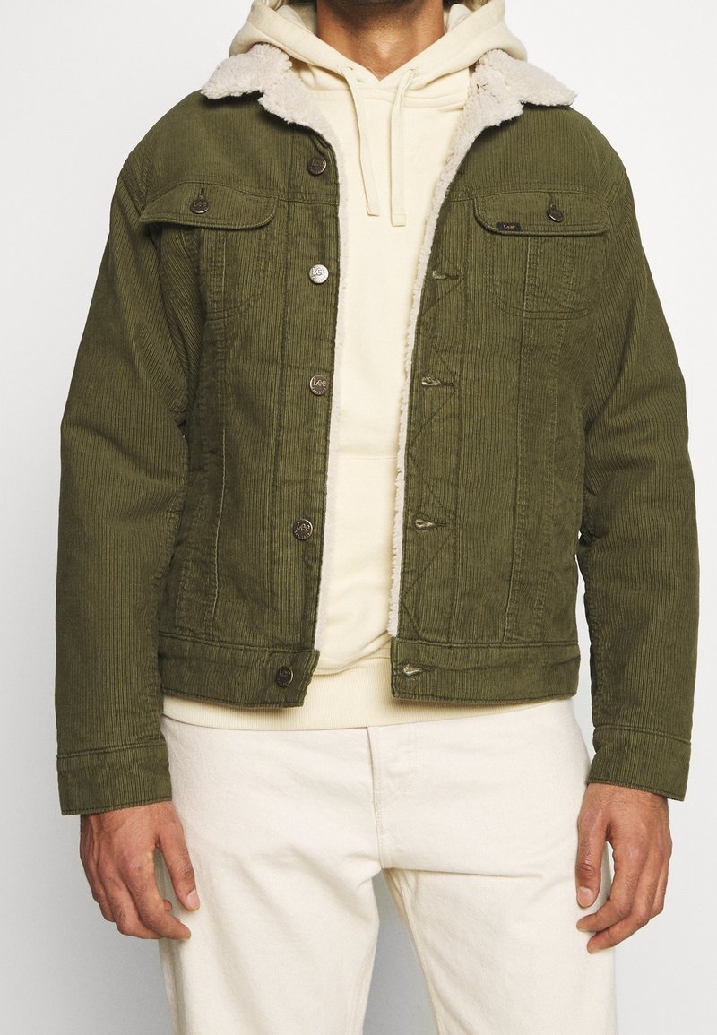 Giacca in corduroy verde oliva con colletto in fascia di shearling finta crema, chiusure a bottoni e tasche sul petto, indossata sopra un leggero hoodie.