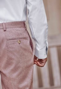 Hellrosa maßgeschneiderte Hose mit glatter Textur, ausgestattet mit einer einzelnen Gesäßtasche und einem Knopfverschluss. Getragen mit einem weißen Hemd.