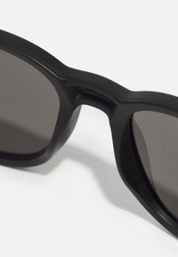 Occhiali da sole neri con una montatura spessa e opaca e lenti scure. Presentano un design del ponte unico e una texture liscia. Nessun marchio visibile.