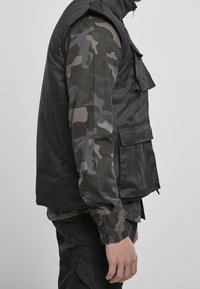 Černá vesta bez rukávů s několika kapsami, nošená přes maskáčovou košili s dlouhým rukávem. Hladká látka s knoflíky a zapínáním na zip u krku.