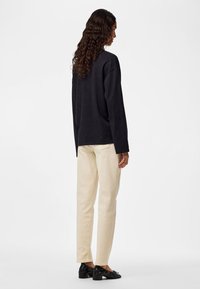 Persona con capelli lunghi e ricci che indossa una maglia nera a maniche lunghe, pantaloni color crema e scarpe nere con tacco basso, in piedi davanti a uno sfondo bianco uniforme.