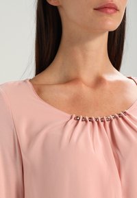 Jasnoróżowa bluzka z okrągłym dekoltem, marszczonym materiałem z przodu i ozdobnymi koralikami wzdłuż dekoltu. Miękka, gładka faktura.