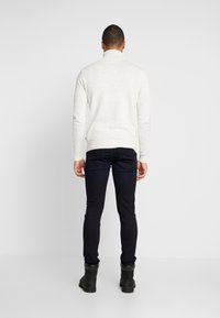Suéter blanco moteado con cuello alto y forma ajustada. Combinado con jeans oscuros de corte slim y botas de tobillo negras. Fondo liso.