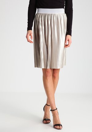 Femme portant une jupe plissée argentée brillante avec une ceinture blanche, un haut noir à manches longues et des sandales à talons noires avec bride à la cheville.