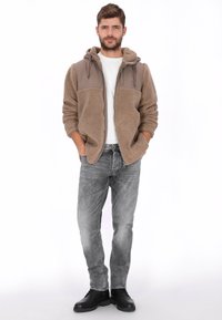 Chaqueta marrón de forro polar con cremallera, parte superior de tela lisa, dos bolsillos delanteros y capucha. Llevada sobre una camiseta blanca y combinada con vaqueros grises y botas negras.