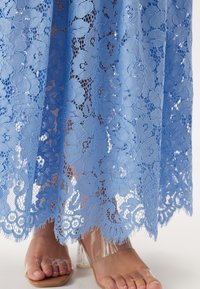 Ljusblå spetsklänning med blommönster, med en scalloped fåll. Transparenta sandaletter synliga längst ner, som visar bara fötter.