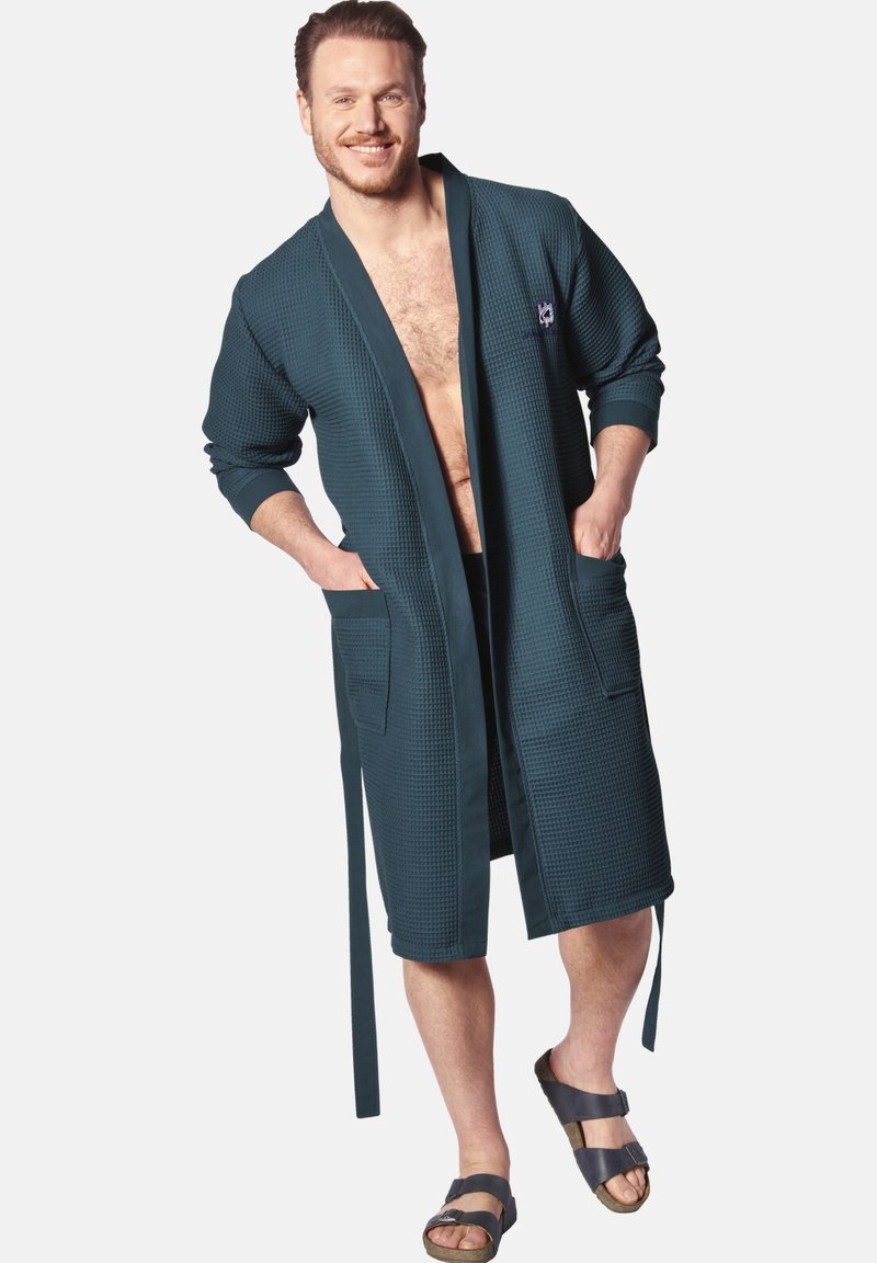 Dark teal waffle-textured bathrobe with long sleeves, two front pockets, and a tie waist. Worn open over shorts, paired with sandals.