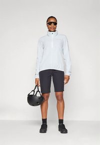 Gaiši zils vējdēlis ar augstu apkakli, melnas šorti un melna ķivere rokā. Modelis stāv taisni, valkājot saulesbrilles.