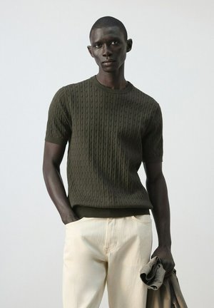 Hombre vistiendo un suéter de punto de manga corta verde oscuro y pantalones color crema, sosteniendo una chaqueta beige doblada en una mano sobre un fondo liso.