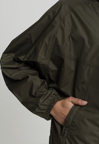 Olivgrön nylonjacka med avslappnad passform, elastiska ärmslut och sidofickor. Slät yta med minimala sömnadsdetaljer.