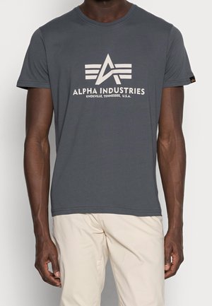 Mężczyzna nosi ciemnoszarą koszulkę Alpha Industries z białym logo i nadrukiem, w połączeniu z jasnobeżowymi spodniami, stoi na jednolitym tle.