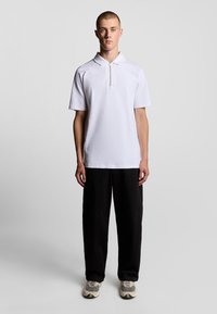 Weiße Poloshirt mit Reißverschlusskragen, aus weichem Stoff. Kombiniert mit lockeren schwarzen Hosen und neutraltonigen Sneakers.