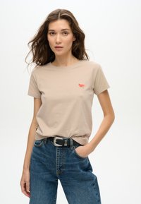T-shirt en coton beige à manches courtes et col rond, avec un petit logo rouge brodé sur la poitrine, associé à un jean taille haute bleu.